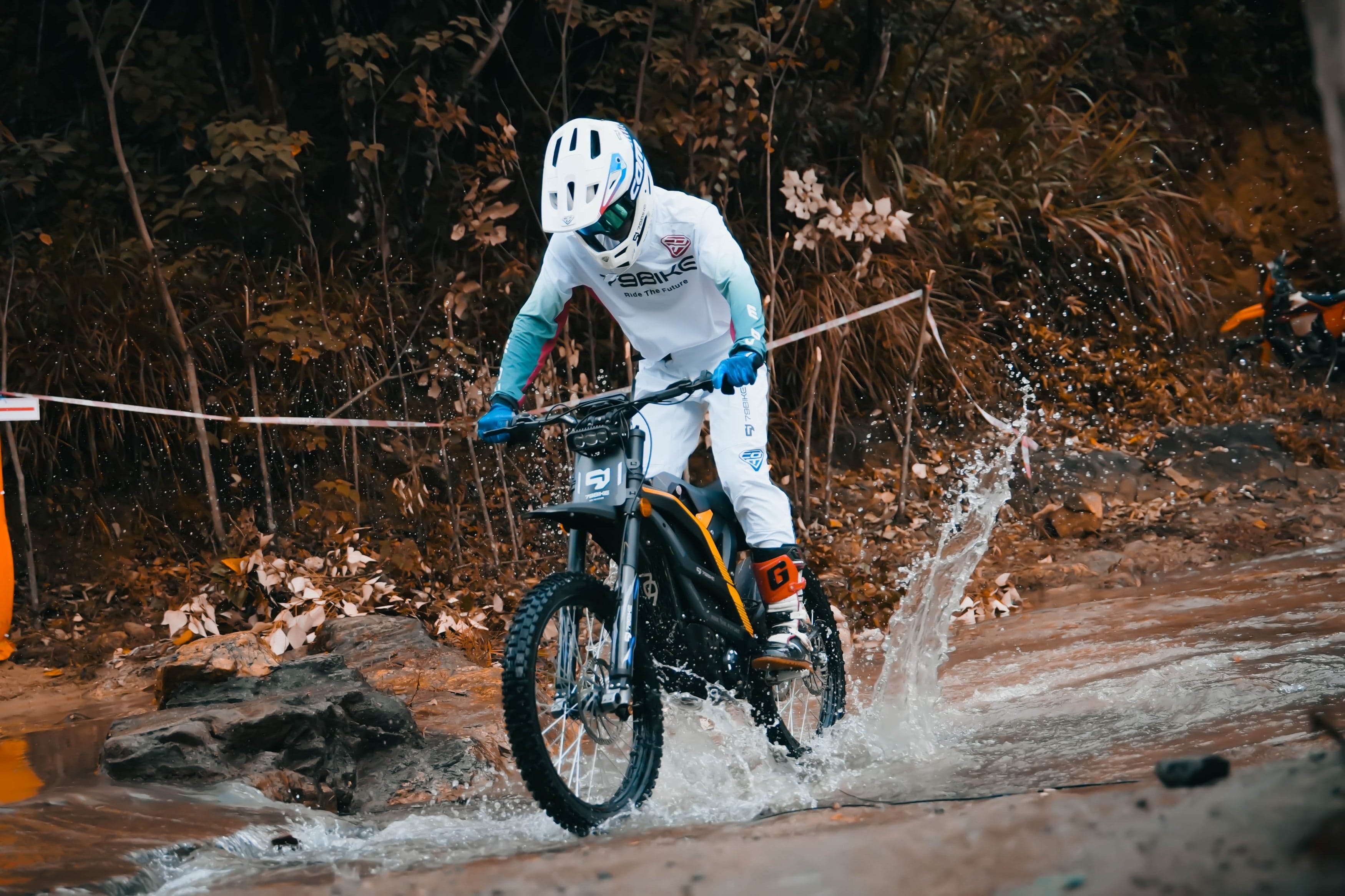 Rider on an electric dirt bike splashing through a shallow creek on a wooded trail